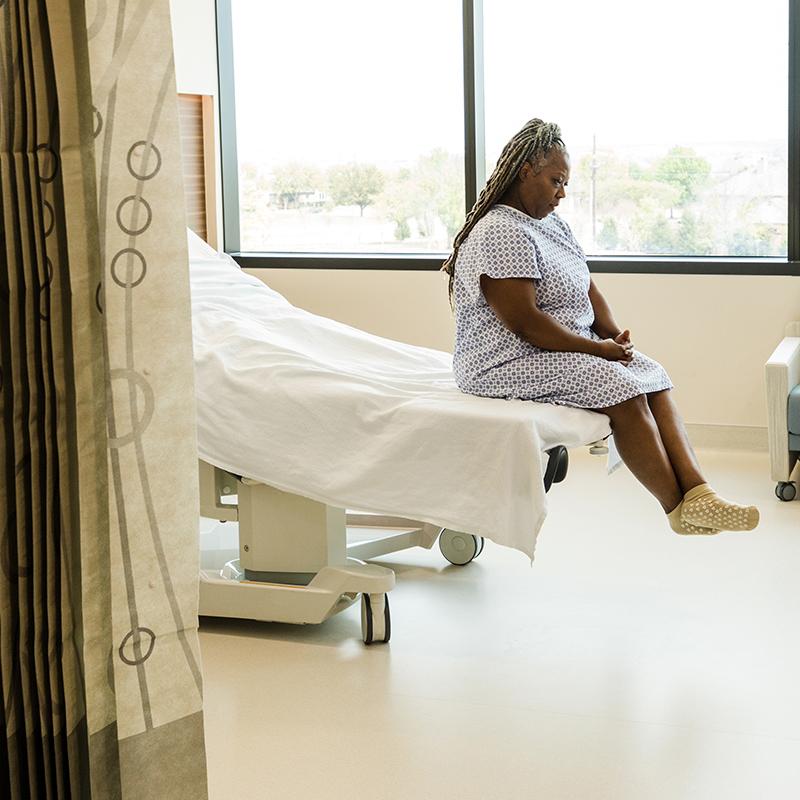 Black woman in a hospital gown sitting on a clinic bed and looking downward
