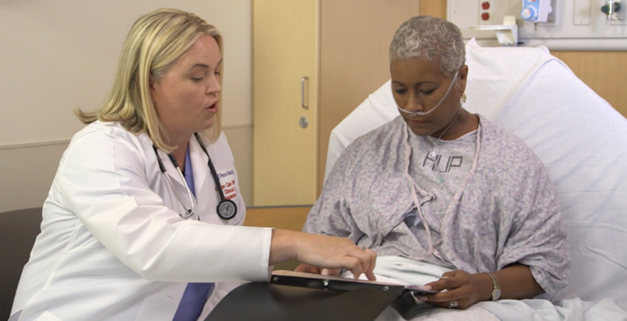 Colleen talking to a patient in a hospital bed beside her and pointing to a paper in the patient's lap