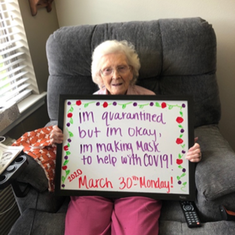 Elderly woman holding a sign saying "Im quarantined but im okay, Im making Mask to help with COV19! 2020 March 20th-Monday!"