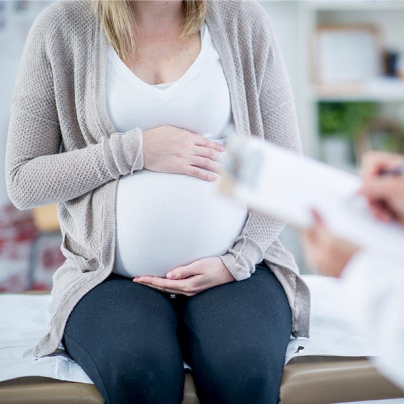 Pregnant woman sitting in an exam room and holding her belly. In the foreground, a doctor writes on a clipboard.