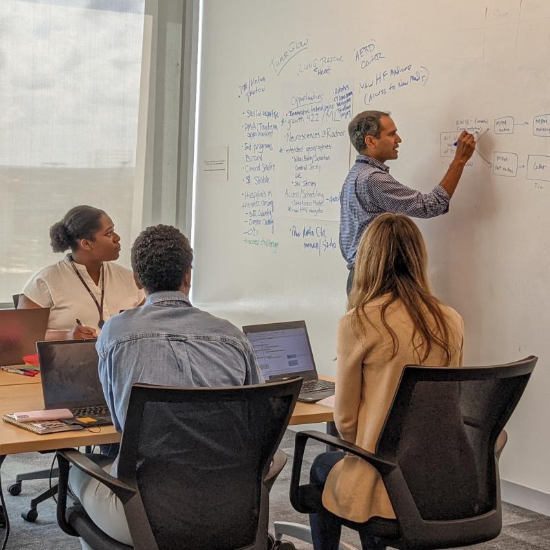 Shivan draws a diagram on a white board in front of team members seated at a table