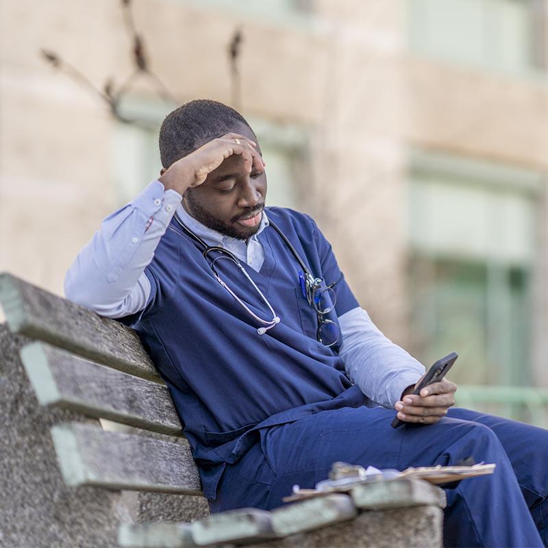 Clinician sitting outside on a bench looking at his phone and with one hand resting on his forehead