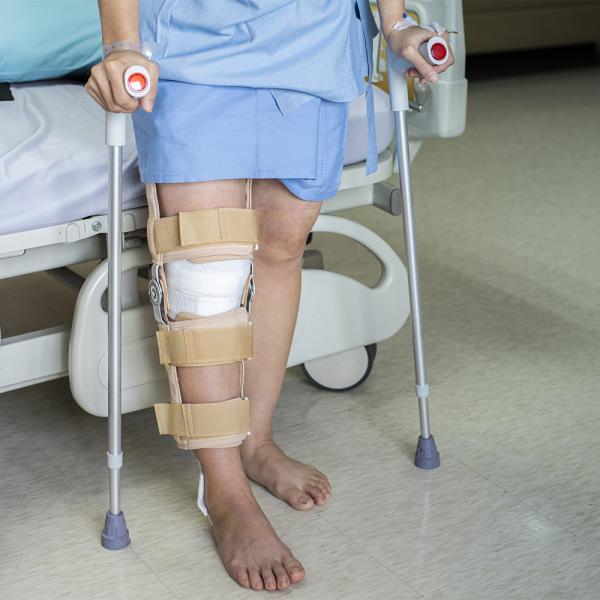 Waist-down shot of someone with their leg in a brace holding canes and standing next to a hospital bed